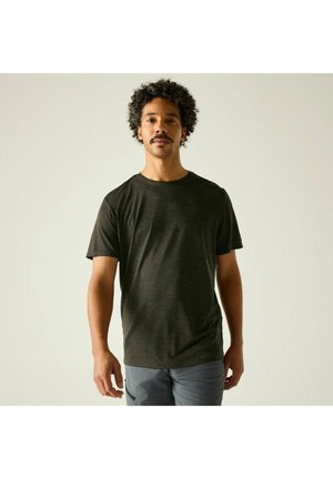 Mężczyzna z kręconymi włosami i wąsami, ubrany w ciemnozielony t-shirt z krótkim rękawem i szare spodnie, stoi na tle jednolitego jasnego tła.