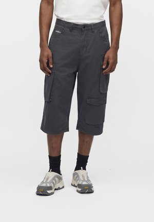 Férfi sötétszürke cargo rövidnadrágot, fehér tornacipőt, fekete zoknit és fehér inget visel, egyszerű háttér előtt áll.