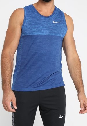 Man wearing a blue sleeveless athletic top and black Nike pants, standing with arms relaxed by his sides.