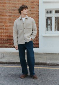 Light beige textured shirt jacket with two pockets, worn over a white t-shirt, paired with dark blue straight-leg jeans and brown shoes.