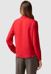 Blouse rouge à manches longues avec une texture douce et lisse, dotée d'un col rond et de poignets boutonnés. La vue de dos révèle un design épuré et ajusté.