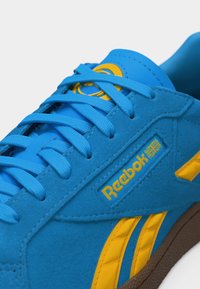 Modri semiš tekaški čevelj Reebok z rumenim logotipom in črtami, modrimi vezalkami ter rjavim podplatom, prikazan v podrobnem posnetku.