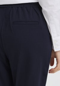 Pantaloni bleumarin cu talie elastică, având un buzunar plat în spate. Textură netedă, fără modele sau elemente vizibile.