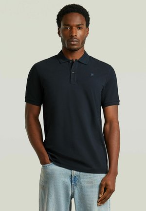 Mann trägt ein schwarzes, kurzärmeliges Poloshirt und hellblaue Jeans, steht mit einer Hand in der Tasche vor einem schlichten Hintergrund.