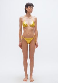 Λεπτή γυναίκα με κοντά σκουρόχρωμα μαλλιά στέκεται ξυπόλυτη, φορώντας ένα λαμπερό χρυσό μπικίνι μπροστά σε ένα απλό λευκό φόντο.