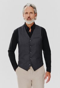 Szary kamizelka o fakturowanym wykończeniu, z pięcioma guzikami i dwiema przednimi kieszeniami, noszona na czarnej koszuli i zestawiona z jasnymi spodniami.