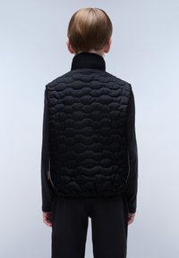 Černá péřová vesta s prošívanou strukturou, vysokým límcem a elastickým lemem. Nosená přes černou košili s dlouhým rukávem, pohled zezadu.