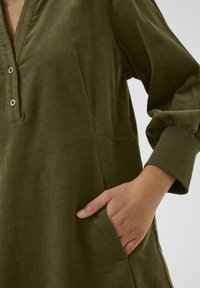 Olive green corduroy tunic with a V-neck, front buttons, 3/4 sleeves, and side pockets. Textured fabric with ribbed accents.