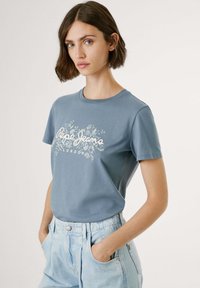 Zila kokvilnas T-krekls ar īsām piedurknēm, kas apvieno ziedu dizainu un baltu tekstu "Pepe Jeans London." Nēsāts ar gaiši ziliem augstiem džinsiem.