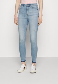 Jeans skinny de tiro alto en azul claro con un leve desgaste y pequeñas roturas. Dobladillos acampanados y combinados con zapatillas blancas con acentos en burdeos.