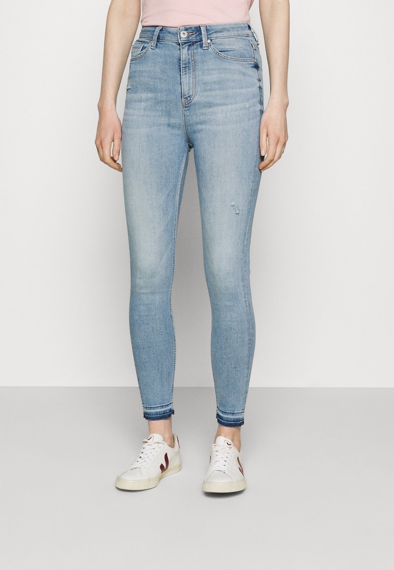 Jeans skinny de tiro alto en azul claro con un leve desgaste y pequeñas roturas. Dobladillos acampanados y combinados con zapatillas blancas con acentos en burdeos.