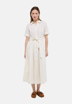 Femme portant une robe midi en dentelle œillet blanche avec manches courtes, col, boutons sur le devant et ceinture nouée à la taille, associée à des mocassins marron.