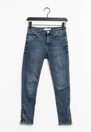 Blauwe skinny jeans van denim met voor- en achterzakken, ritssluitingen aan de onderkant, op een zwarte hanger gehangen tegen een witte achtergrond.