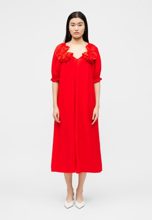 Žena s dlhými čiernymi vlasmi oblečená v voľných červených midi šatách s riaseným výstrihom a nadýchanými rukávmi, stojaca pred jednoliatym svetlým pozadím.