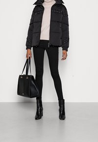 Giubbotto puffo nero con chiusura a zip, spalle testurizzate e cappuccio con cordino. Indossato sopra un maglione beige chiaro, abbinato a jeans skinny neri e stivali.