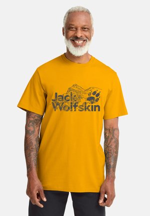 Uomo anziano sorridente con i capelli e la barba bianchi che indossa una maglietta gialla Jack Wolfskin con grafiche di montagna e impronta di zampa.