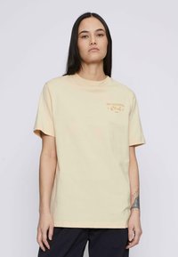 Camiseta de algodón color amarillo claro con mangas cortas, escote redondo y la impresión "Club de No Hacer Nada" en naranja en la parte izquierda del pecho.