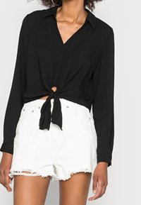 Schwarze Bluse mit langen Ärmeln, V-Ausschnitt und Knoten-Detail vorne, kombiniert mit weißen, verwaschenen Jeansshorts mit fransigen Säumen.