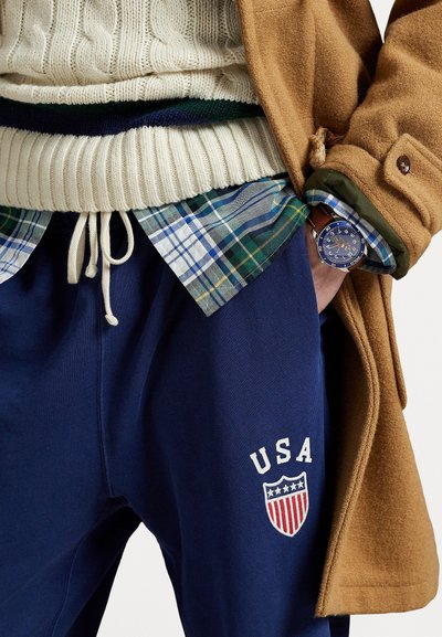 Pantaloni de trening navy cu inscripția "USA" și emblemă în formă de scut, asortați cu un pulover din lână cu model împletit de culoare crem și o cămașă flanelată cu carouri. Paltonul maro este expus.