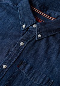 Camisa de mezclilla en azul oscuro con cuello abotonado, botones blancos, bolsillo en el pecho y etiqueta de la marca cosida en rojo en el interior del cuello.