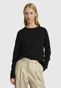 Černý dlouhý rukáv top v uvolněném střihu, vyrobený z měkké tkaniny. V kombinaci s béžovými skládacími kalhotami, ukazující jednoduchý výstřih a čisté linie.
