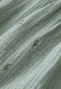 Tissu vert clair à surface texturée, présentant des motifs botaniques brodés subtils et de petits accents blancs répartis sur l'ensemble.