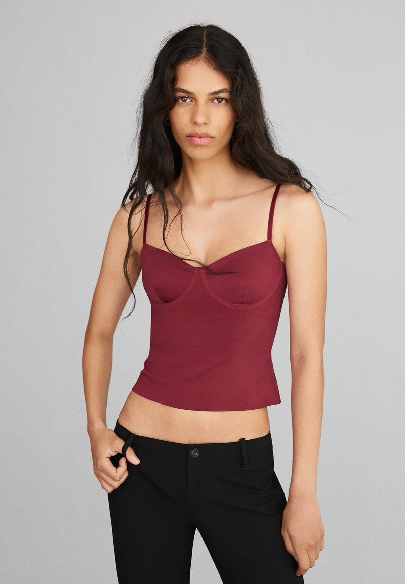 Femme aux longs cheveux foncés portant un top caraco bordeaux ajusté et un pantalon noir sur un fond gris uni.