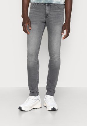 Homme portant un jean gris slim et des baskets blanches, debout devant un fond clair uni.