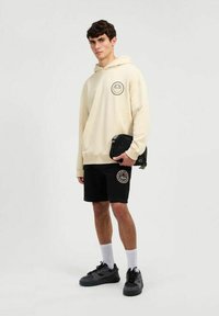 Sudadera beige con un logo circular, pantalones cortos negros con un logo, zapatillas negras y calcetines blancos, de pie frente a un fondo neutral.
