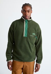 Man with braided hair wearing a green Columbia fleece pullover with snap buttons and brown pants, standing against a plain white background.