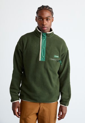 Mann mit geflochtenem Haar, der einen grünen Columbia-Fleece-Pullover mit Druckknöpfen und braunen Hosen trägt, steht vor einem schlichten weißen Hintergrund.