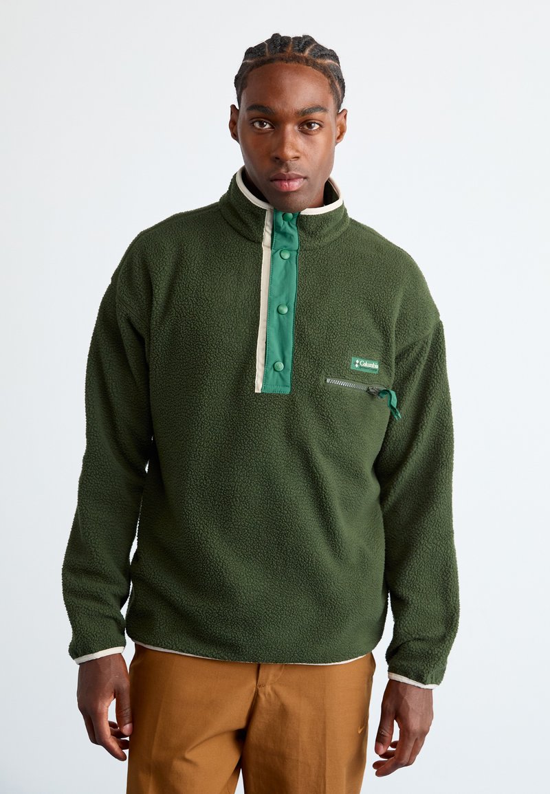Man with braided hair wearing a green Columbia fleece pullover with snap buttons and brown pants, standing against a plain white background.