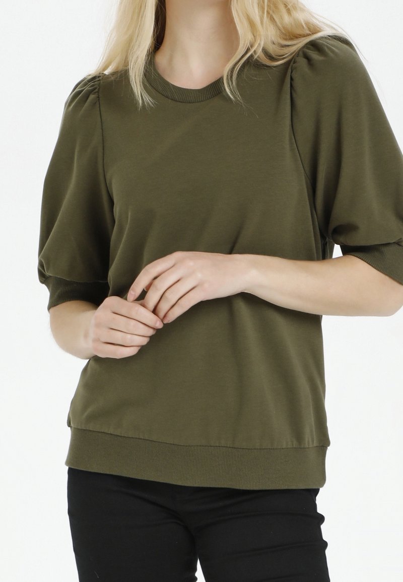 Femme portant un haut vert olive à manches courtes bouffantes, avec encolure et ourlet côtelés, debout les mains jointes doucement.