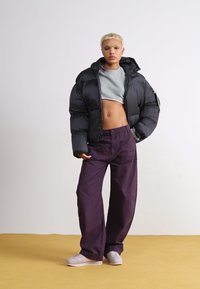 Zwarte gewatteerde jas, grijze cropped top en losse donkerpaarse broek. Model draagt lichte sneakers tegen een minimalistische achtergrond.