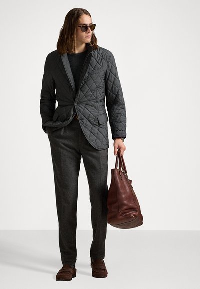 Mann in dunkler gesteppter Blazer, grauen Hosen, braunen Slippern, Sonnenbrille, der mit einer Hand eine große braune Lederhandtasche hält und vor einem weißen Hintergrund steht.