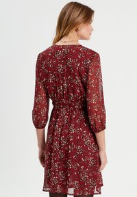 Robe florale bordeaux avec des manches transparentes trois-quarts, taille smockée, tissu léger et une silhouette fluide et lisse.