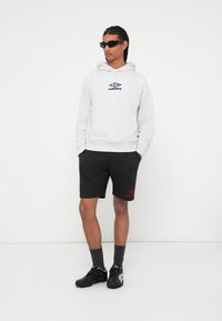 Sudadera gris con capucha y logo, pantalones cortos negros con un logo rojo, zapatillas deportivas negras y gafas de sol. Apariencia casual y deportiva.