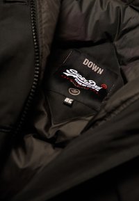Svart dunjacka inuti med en etikett som har "Superdry"-logotyp, som anger "DOWN", med en slät, matt tygstruktur.