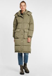 Olivgrön pufferjacka med knälång design, framknäppning, två stora framfickor och avtagbar huva, tillsammans med blå jeans.