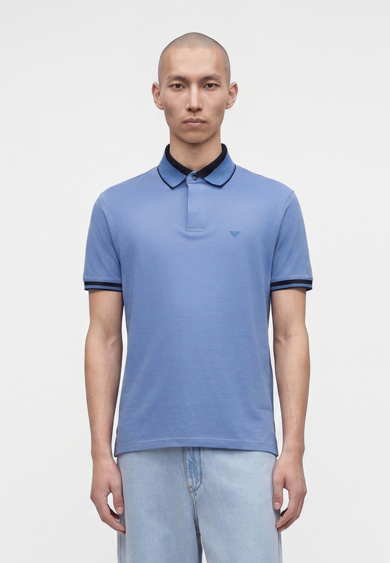 Άνδρας με ξυρισμένο κεφάλι, φορώντας μπλε polo κοντομάνικο με σκούρο τελείωμα και ανοιχτό μπλε τζιν, στέκεται μπροστά σε απλό λευκό φόντο.