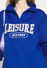 Blå sweatshirt med krage, med "LEISURE" och "1998" i vit och orange text över framsidan. Mjuk textilmaterial.