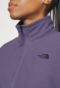 Fleecowa kurtka w kolorze purpurowym z wysokim kołnierzem, zapięciem na zamek oraz czarnym haftowanym logo po lewej stronie klatki piersiowej. Gładka, miękka tekstura.