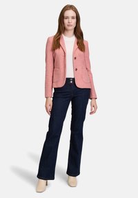 Valkoinainen bleiseri, jossa on kaksi etutaskua, yksinappinen kiinnitys ja kuvioitu kangas, yhdistetty tummansiniisiin liehukehousuihin ja paljaisiin korkokenkiin.