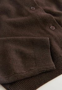 Brun cardigan med en blød, tekstureret strik. Har to brune knapper og ribbede manchetter. Varmt stof der egner sig til lag-på-lag.