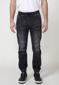 Joggers de denim negro con cintura y puños elásticos, que cuentan con costuras en capas y bolsillos frontales. Textura suave con un acabado desgastado.