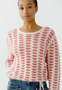 Woman wearing a white fuzzy sweater patterned with red lips paired with blue denim jeans, standing against a plain light background.