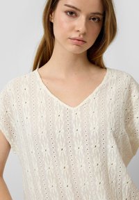 Joven con cabello liso castaño claro, vestida con una blusa de punto texturizada color crema, de escote en V y mangas cortas.
