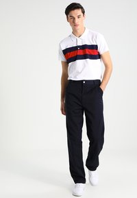Witte polo met rood en marineblauwe horizontale strepen, gecombineerd met marineblauwe broek en witte sneakers. Glad materiaal, casual pasvorm en knoopdetails.