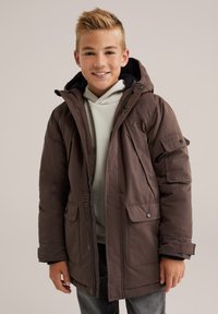 Nasmejan deček oblečen v rjavo zimsko jakno s kapuco, pod katero ima svetlo bež kapuco, ter temne hlače pred enobarvno ozadje.