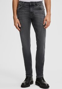 Uomo che indossa jeans neri slim-fit sbiaditi e stivali in pelle neri con lacci, in piedi contro uno sfondo grigio chiaro semplice.